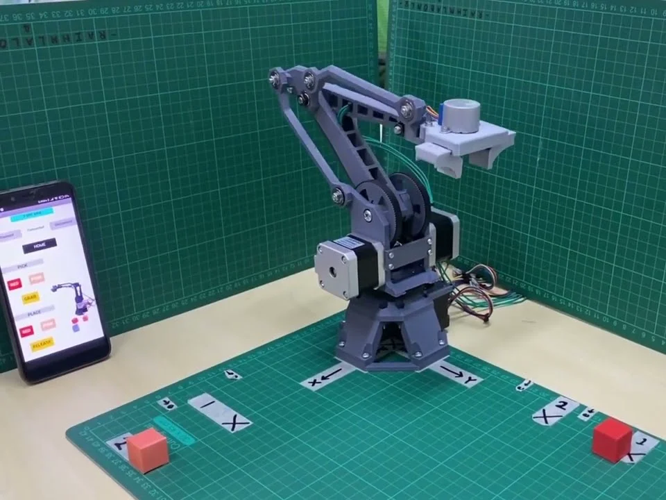 The three-jointed arm on a cutting mat, belt-driven with stepper motors at each joint and a gripper at the end, with coloured cubes laid out in front of it
