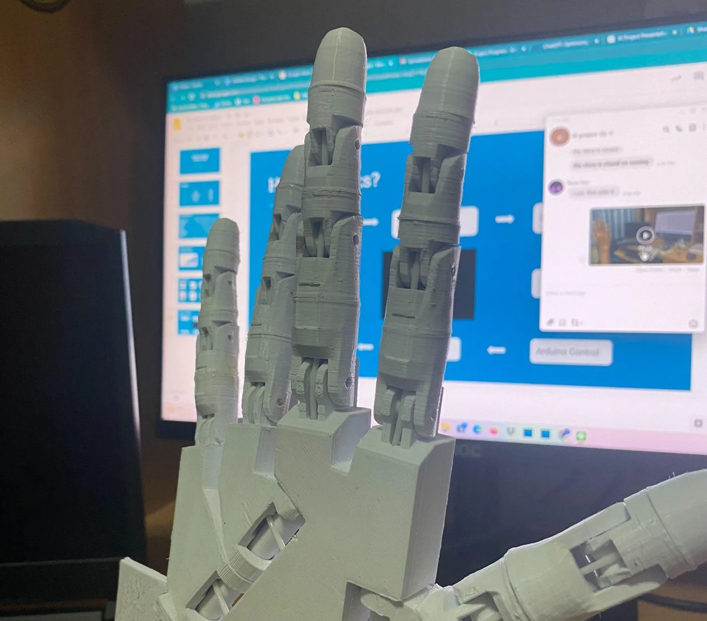 The 3D-printed robotic hand held up in front of a monitor, fingers extended, servo cables running from the wrist