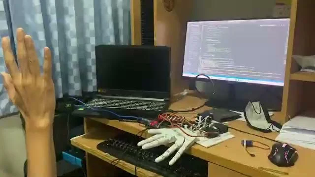 The workspace during a test: a hand raised in front of the webcam on the left, the printed hand and its wiring on the desk, and the code running on the monitor behind