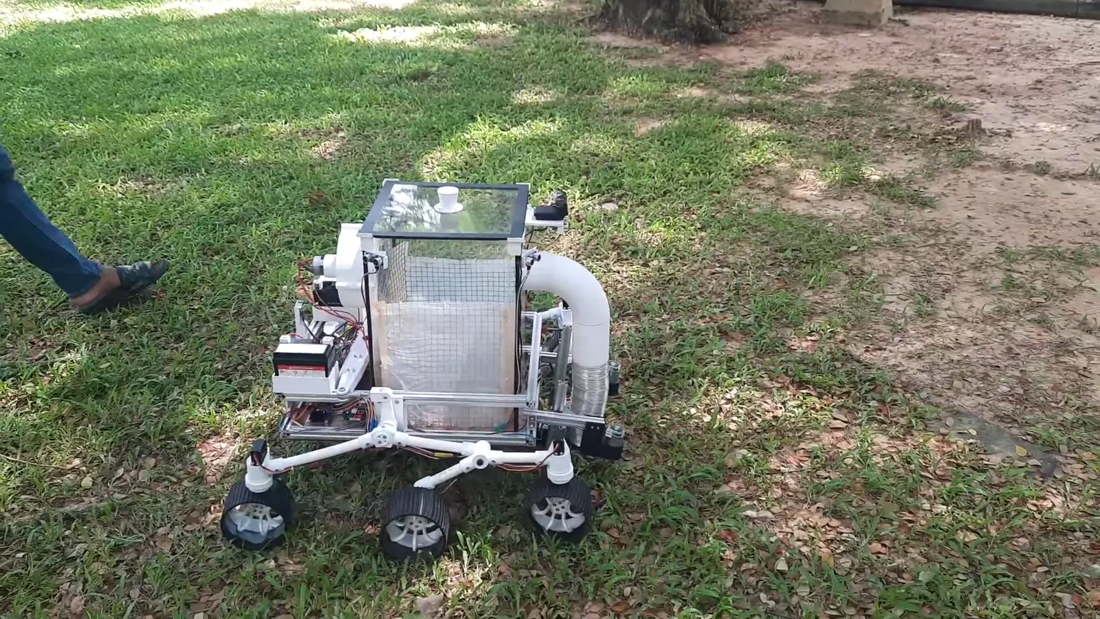 The Leave Collector Robot, a six-wheeled rocker-bogie rover with a vacuum duct and a mesh collection box, driving across grass